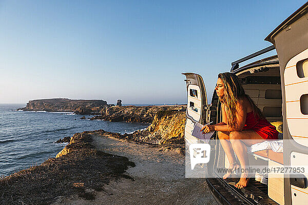 Junge Frau mit Buch bewundert die Aussicht  während sie im Wohnmobil am Strand sitzt