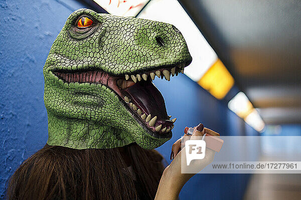 Frau trägt Lippenstift auf  während sie eine Dinosauriermaske vor einer blauen Wand trägt