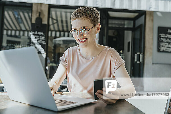 Lächelnde Frau mit Laptop und Smartphone in einem Straßencafé an einem sonnigen Tag