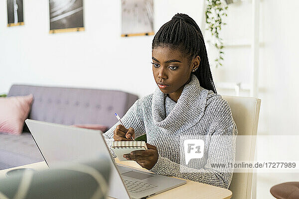 Studentin schreibt in einen Notizblock  während sie zu Hause am Laptop lernt