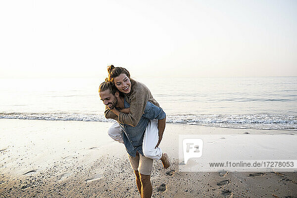 Happy man giving piggyback to girlfriend while walking on shore at beach against clear sky during sunset