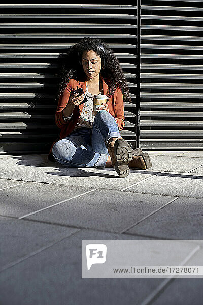 Frau  die ein Smartphone benutzt und eine Kaffeetasse hält  während sie gegen einen Fensterladen sitzt