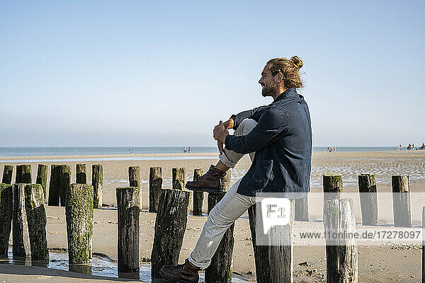 Junger Mann betrachtet die Aussicht  während er auf einem Holzpfosten am Strand gegen den klaren Himmel sitzt