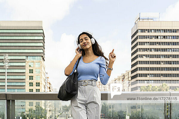 Junge Geschäftsfrau genießt Musik auf Kopfhörern  während sie sich auf einem Geländer gegen den Himmel lehnt