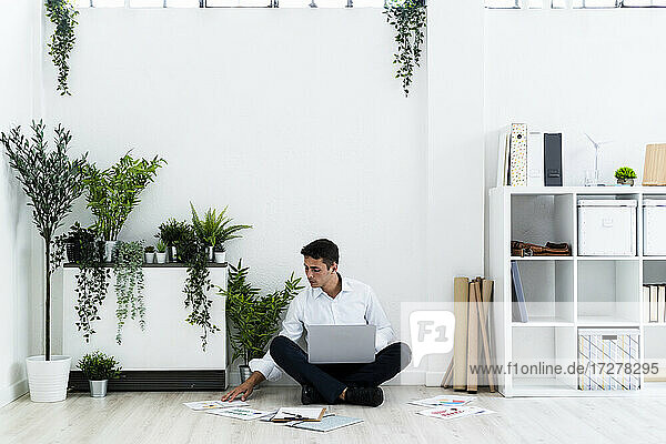 Junger Geschäftsmann  der Dokumente analysiert  während er mit seinem Laptop auf dem Boden vor einer weißen Wand im Büro sitzt
