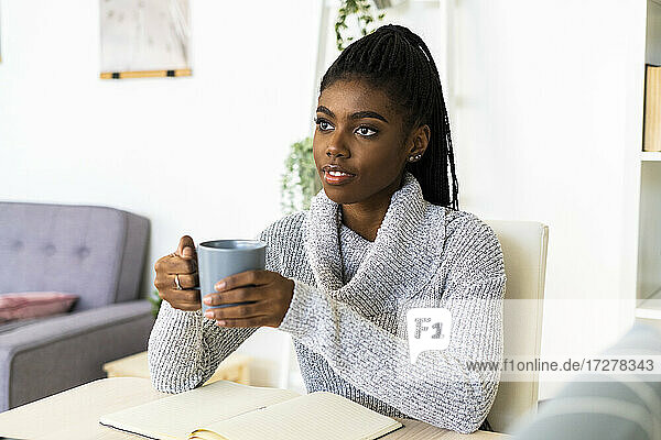 Nachdenkliche Frau hält Kaffeetasse und schaut weg  während sie zu Hause im Wohnzimmer sitzt