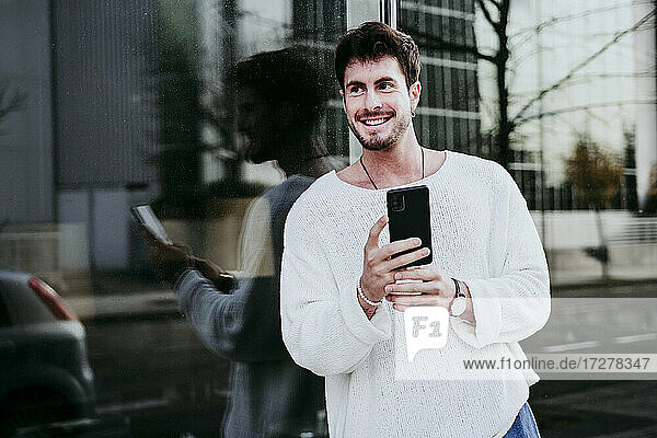 Smiling man with smart phone looking away against building in city