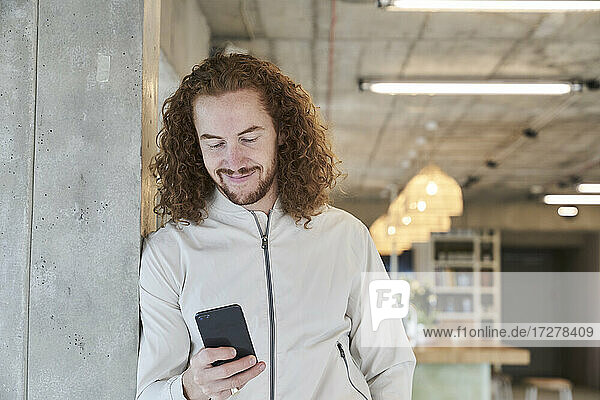 Lächelnder rothaariger Mann  der ein Smartphone benutzt und sich zu Hause an eine Säule lehnt