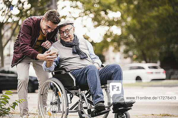 Älterer Mann im Rollstuhl und sein erwachsener Enkel schauen gemeinsam auf ein Smartphone
