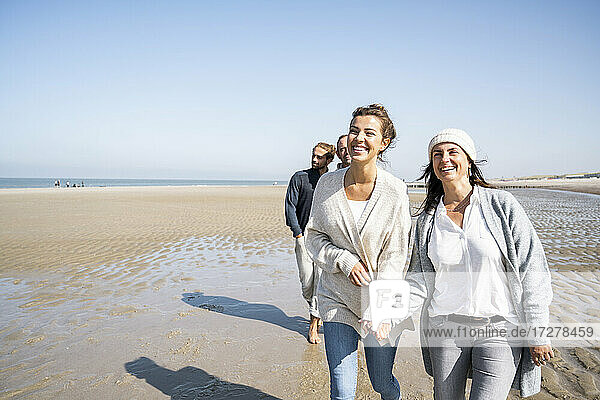 Lächelnde Frauen halten sich an den Händen  während sie mit Männern im Hintergrund am Strand spazieren gehen