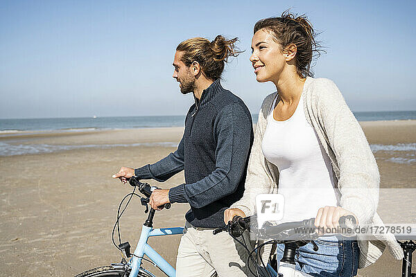 Smiling young woman walking with boyfriend and bicycles at beach against clear sky on sunny day