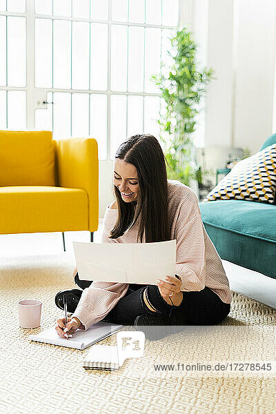 Lächelnde junge Frau  die in einen Notizblock schreibt  während sie mit einer Kaffeetasse auf dem Teppich vor dem Sofa in einer Loftwohnung sitzt