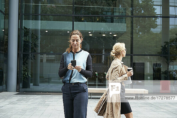 Geschäftsfrau  die ein Mobiltelefon benutzt  mit einem Kollegen  der nach der Arbeit geht  im Hintergrund