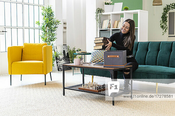 Smiling young woman collecting books in box on coffee table at loft apartment