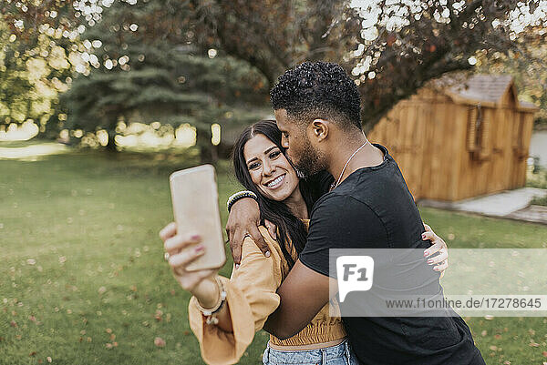 Mann küsst Frau  die ein Selfie mit ihrem Mobiltelefon macht  während sie im Hinterhof steht