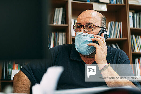 Businessman wearing protective face mask while talking on smart phone in office during COVID-19