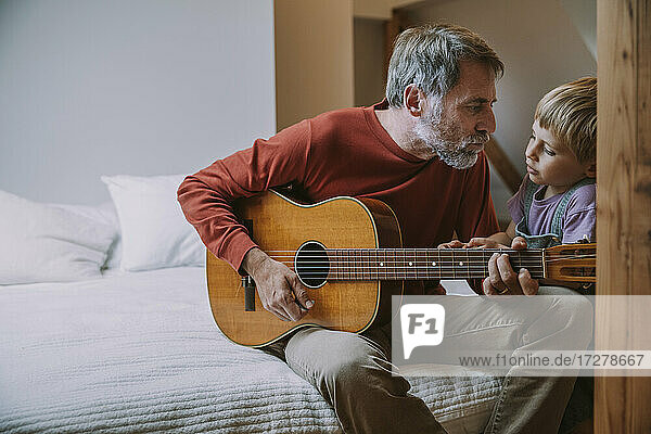 Father playing guitar in front of son while sitting on bed at home