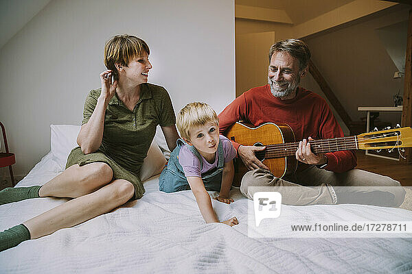 Happy father playing guitar while mother and son sitting on bed at home