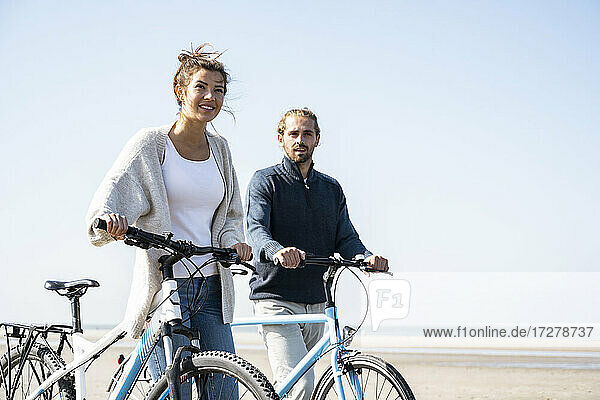 Young couple walking with bicycles at beach against clear sky on sunny day