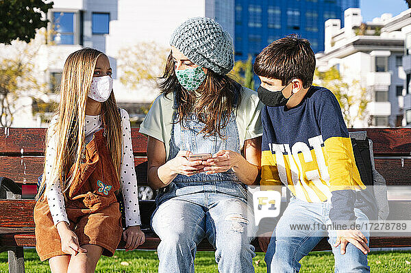 Mutter und Kinder mit Gesichtsschutzmaske und Smartphone auf einer Bank in einem öffentlichen Park an einem sonnigen Tag