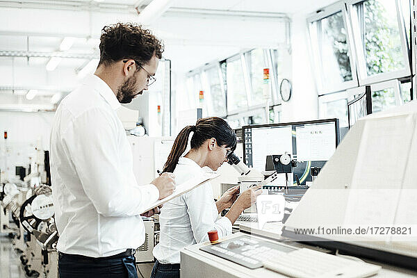 Ein männlicher Techniker schreibt auf einem Klemmbrett  während er neben einer weiblichen Kollegin steht und durch ein Mikroskop im Labor schaut.