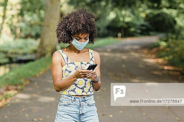 Mittlere erwachsene Frau mit Gesichtsmaske  die ein Mobiltelefon benutzt  während sie auf der Straße im Park steht