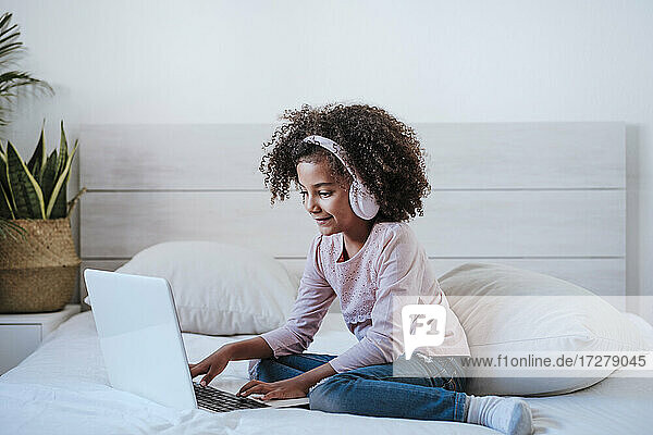 Nettes Mädchen  das einen Laptop benutzt und Kopfhörer trägt  während es im Schlafzimmer zu Hause sitzt
