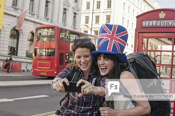 Happy woman taking selfie with friend wearing British flag hat against red telephone box in city