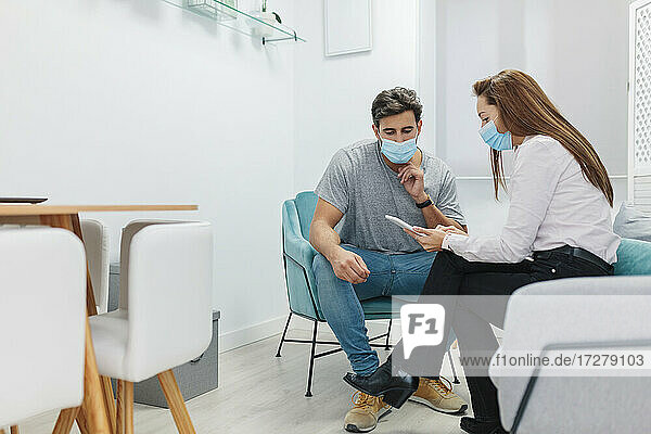 Arzt und Patient tragen eine Gesichtsmaske und benutzen ein digitales Tablet  während sie im Büro sitzen