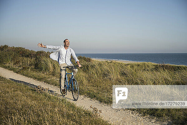 Carefree mature man riding bicycle at beach against clear sky during weekend