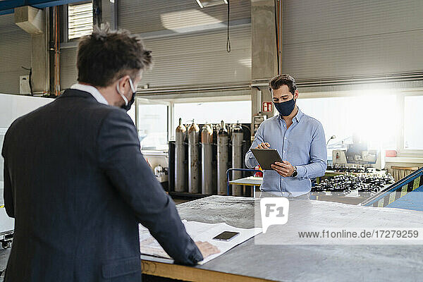 Young businessman with digital tablet and face mask working while standing with colleague at social distance in factory