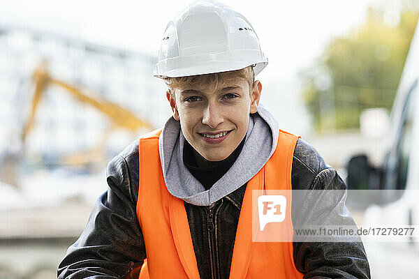 Lächelnder jugendlicher Baupraktikant mit Schutzhelm und reflektierender Kleidung auf der Baustelle
