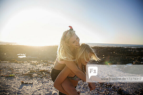 Young woman piggybacking cheerful female friend at beach during sunset