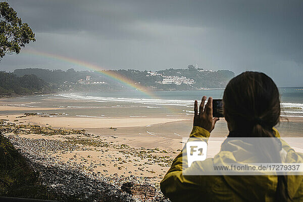 Rückansicht einer Frau im Regenmantel  die einen Regenbogen am Strand fotografiert