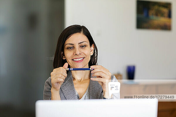 Smiling woman looking at pen while using laptop at home