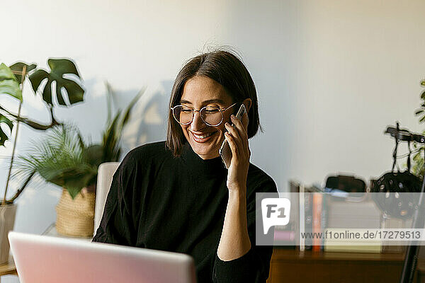 Smiling businesswoman talking on smart phone while using laptop sitting at home