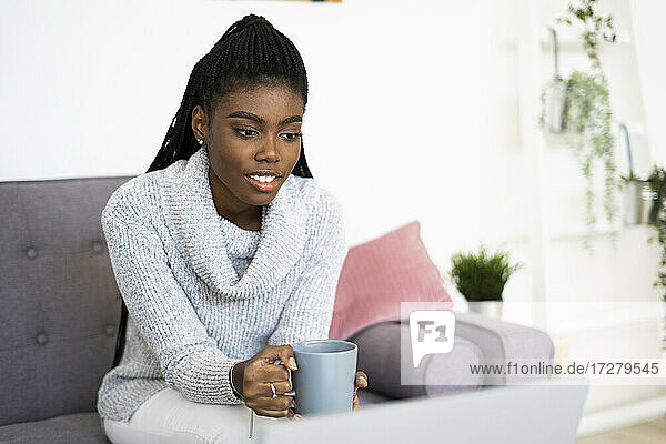 Junge Unternehmerin hält eine Kaffeetasse  während sie zu Hause am Laptop arbeitet