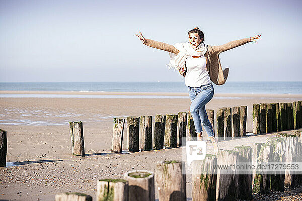 Fröhliche junge Frau mit ausgestreckten Armen  die an einem sonnigen Tag zwischen Holzpfählen am Strand läuft