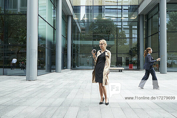 Woman drinking coffee with businesswoman walking in background at city