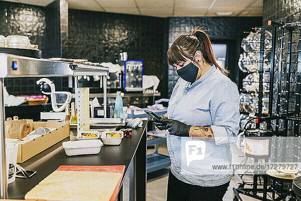 Female chef using smart phone while standing at kitchen counter in restaurant during coronavirus