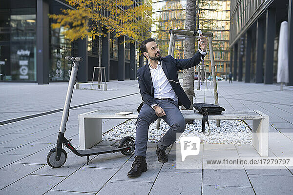 Businessman taking selfie through smart phone by push scooter on bench in city