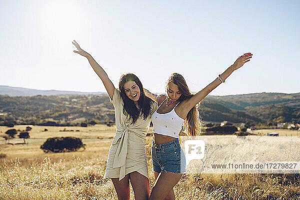 Cheerful young female friends standing with hands raised on grassy field against sky