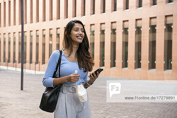 Lächelnde junge Geschäftsfrau mit Smartphone  die einen Gesichtsschutz und ein Händedesinfektionsmittel auf einem Fußweg hält