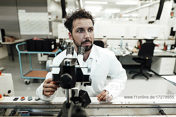 Männlicher Wissenschaftler  der mit einem Mikroskop in einem hellen Labor sitzt und wegschaut