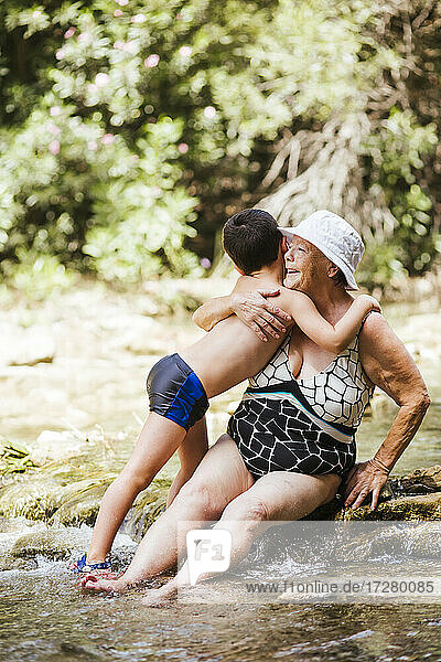 Enkel  der seinen Enkel umarmt  während er auf einem Stein im Fluss im Wald sitzt