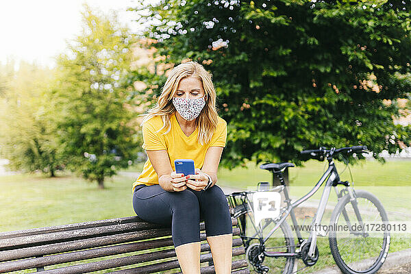 Frau mit Gesichtsschutzmaske und Smartphone auf einer Bank in der Stadt sitzend