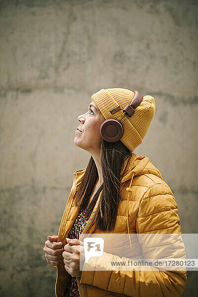 Thoughtful woman in warm clothing listening music through headphones while standing by wall
