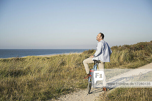 Smiling man looking at view while standing with bicycle on beach against clear sky