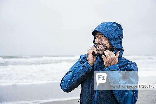 Glücklicher reifer Mann in blauem Regenmantel  der wegschaut  während er am Strand steht
