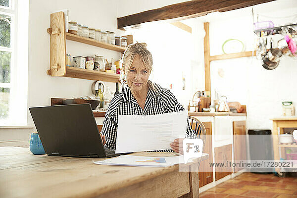 Aktive ältere Frau  die mit ihrem Laptop in der Küche zu Hause sitzt und auf Papier schaut